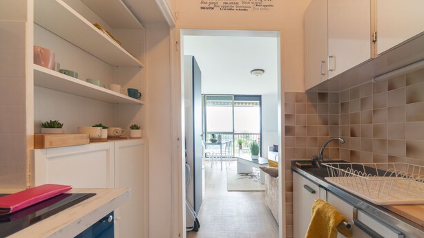 Een smalle keuken met witte kastjes, een spoelbak en een aanrecht. Er is een raam met uitzicht op een balkon en zitplaatsen buiten.