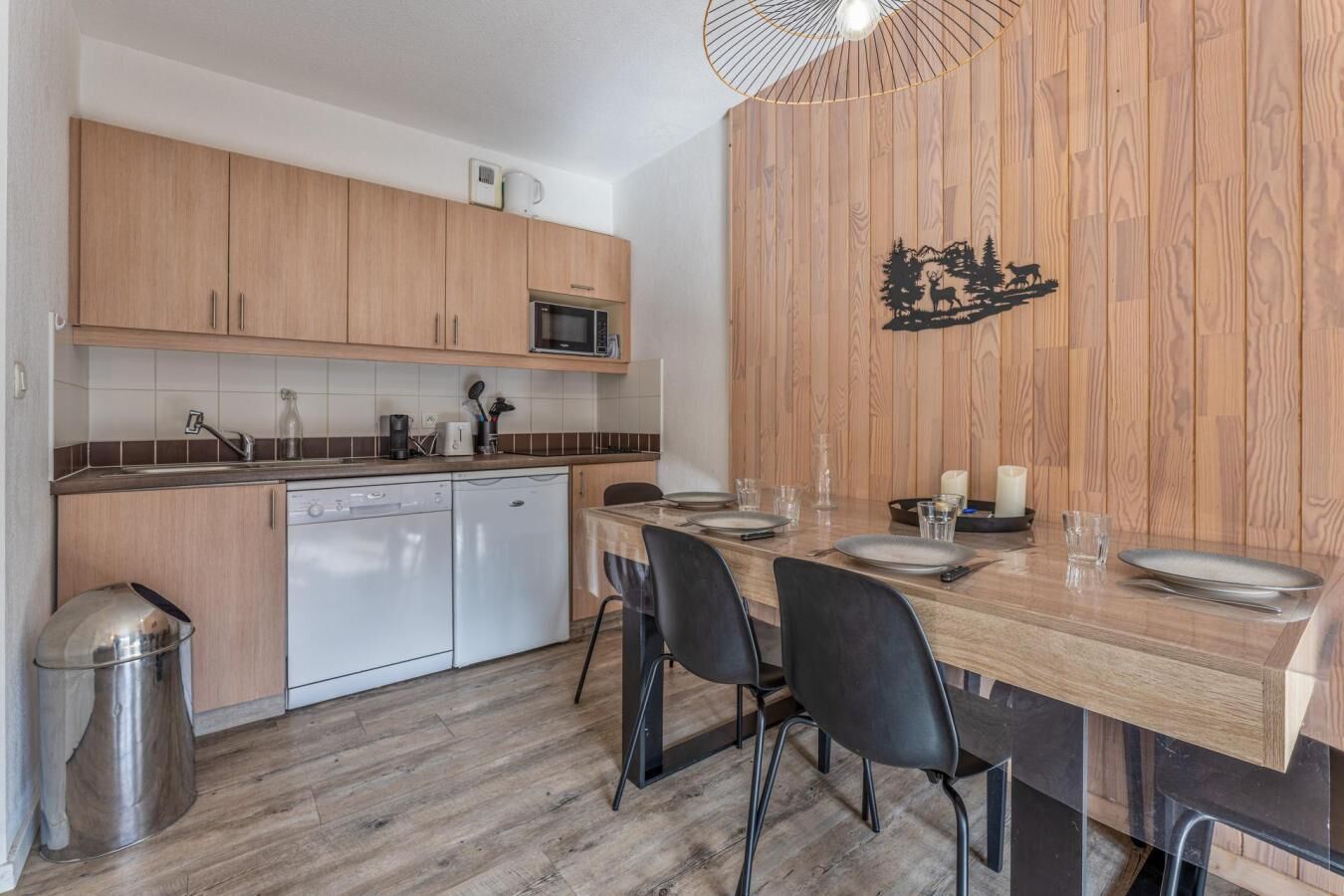 Una cocina compacta con armarios de madera, un microondas y un lavavajillas. Una mesa de comedor con sillas está colocada en el centro.