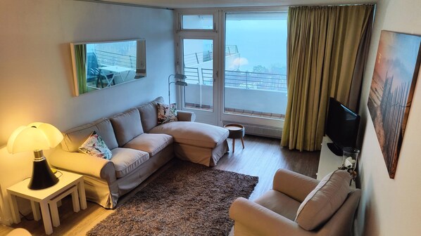 A cozy living room with a sofa, a small table with a lamp, a TV, and a large window with curtains. - Herrlicher Meerblick 911 (Timmendorfer Strand)