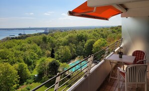 A balcony with a view of a swimming pool, trees, and a distant cityscape. - Herrlicher Meerblick 911 (Timmendorfer Strand)