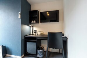 Een kamer met een bureau, stoel, televisie, plank en prullenbak.