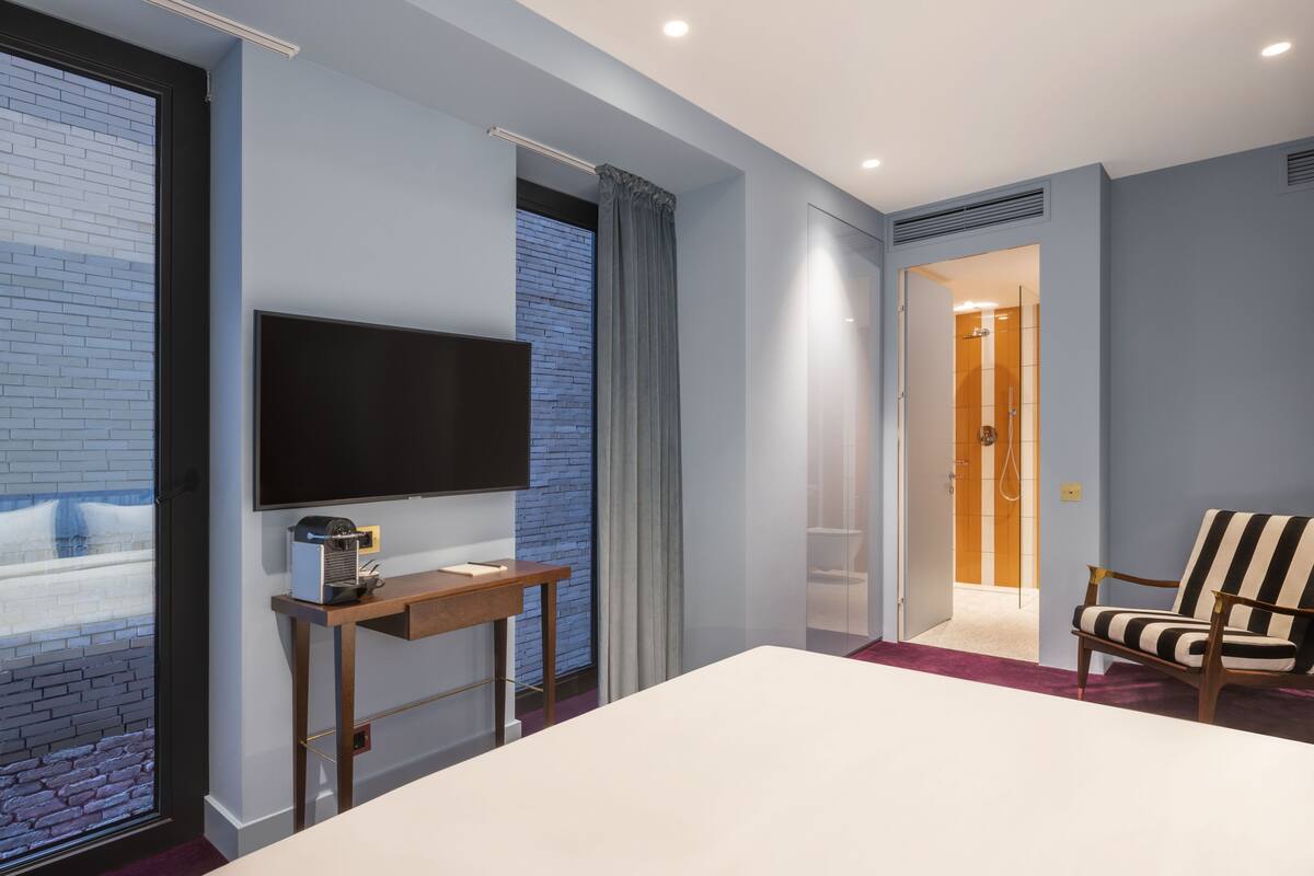 A modern hotel room with a large bed, a flat-screen TV, a desk with a lamp, a striped armchair, and a view through a window with a brick wall.