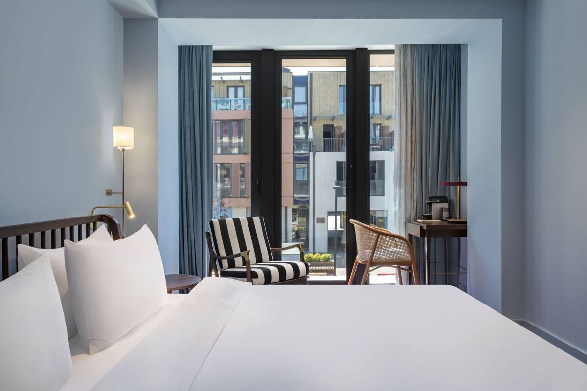 A hotel room with a large bed, two chairs, a desk with a lamp, and a view of buildings through large windows.