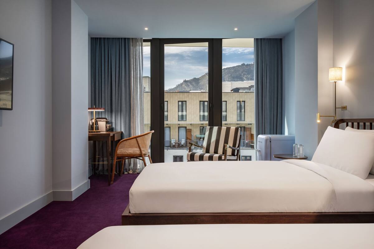A hotel room with a large bed, a desk with a chair, a lamp, and a view of buildings and mountains through the window.