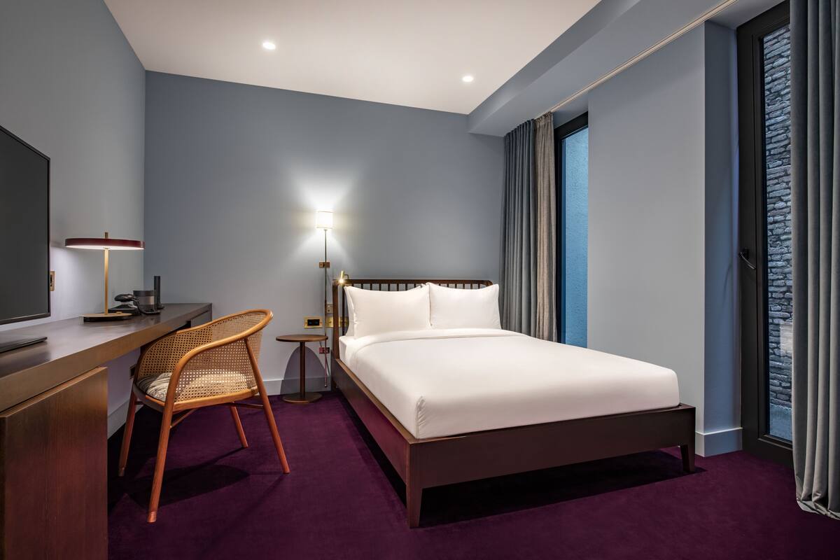 A hotel room with a bed, desk, chair, and a lamp.