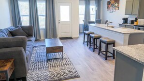 Una sala moderna con sofá, mesa de centro y alfombra estampada. Se ve un área de cocina con una barra y taburetes.