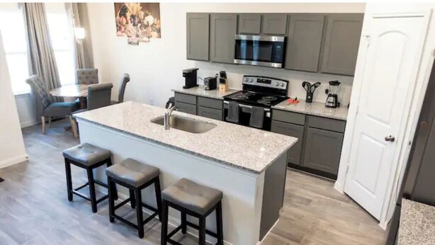 Una cocina moderna con gabinetes grises, encimera de granito, estufa de acero inoxidable y un lavamanos. Hay tres taburetes en la isla.