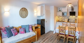 A cozy living area with a sofa, a wooden side table, and a dining table with chairs. A kitchen with a microwave and a stove is visible in the background. - 150m from resort center, 1st floor, view resort, swimming pool, sauna, terrace, ski locker, 33m² (Les Orres)