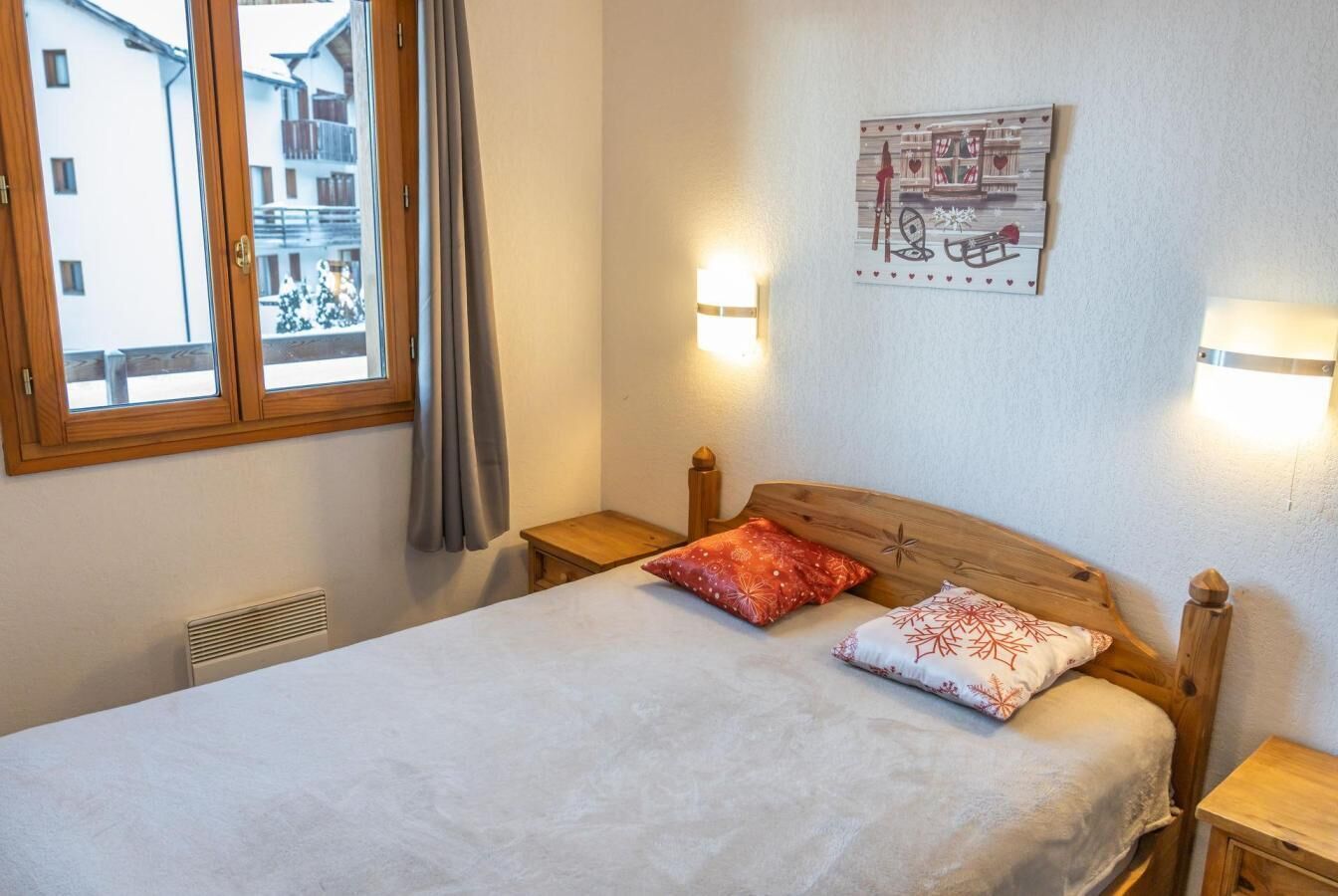 A bedroom with a wooden bed, two bedside tables, a wall-mounted light, and a window with a view of buildings.