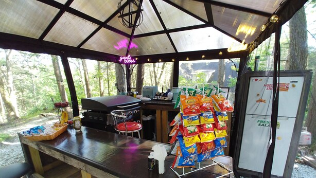 Eine überdachte Außenküche mit Grill, Barhocker und einem mit Snacks gefüllten Kühlschrank.