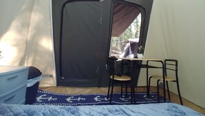 Ein Zelt mit Tisch und Stühlen im Inneren, einem Bett mit blauer Decke und einem Fenster mit Blick auf Bäume.