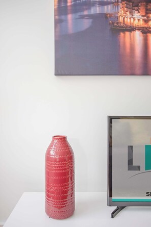 A red vase on a white table, a framed picture with the letters "L" and "S" on a wall, and a small boat in a body of water.