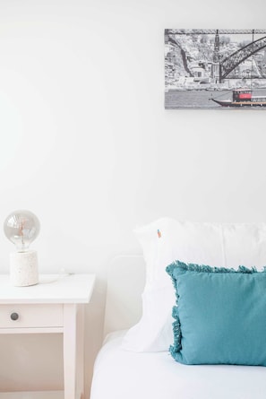 A white bed with a teal pillow, a white nightstand with a lamp, and a black and white cityscape painting on the wall.