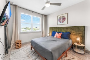 A bedroom with a bed, a TV, a ceiling fan, a window with blinds, a small side table, and a wall art piece.
