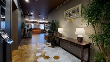 A hotel lobby with a reception desk, a wooden floor with hexagonal tiles, a reception area with a counter and chairs, and a large potted plant.