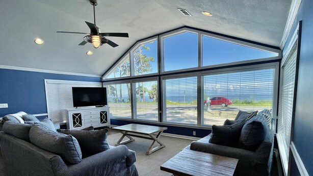 A spacious living room with a large window, a ceiling fan, and a flat-screen TV.