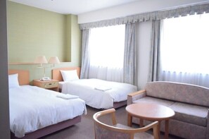 Two single beds with white linens, a nightstand and lamp, and a sofa in a light green room with a large window.