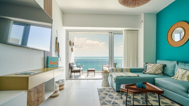 Un salon moderne avec un canapé de couleur turquoise, une table basse et une vue sur l’océan grâce à de grandes fenêtres.