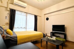 Habitación de hotel con cama, televisor, una mesa con un florero y ventana con cortinas.