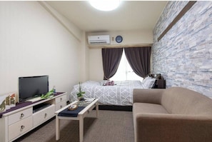 A compact hotel room with a bed, a sofa, a TV, a small table, and an air conditioning unit.