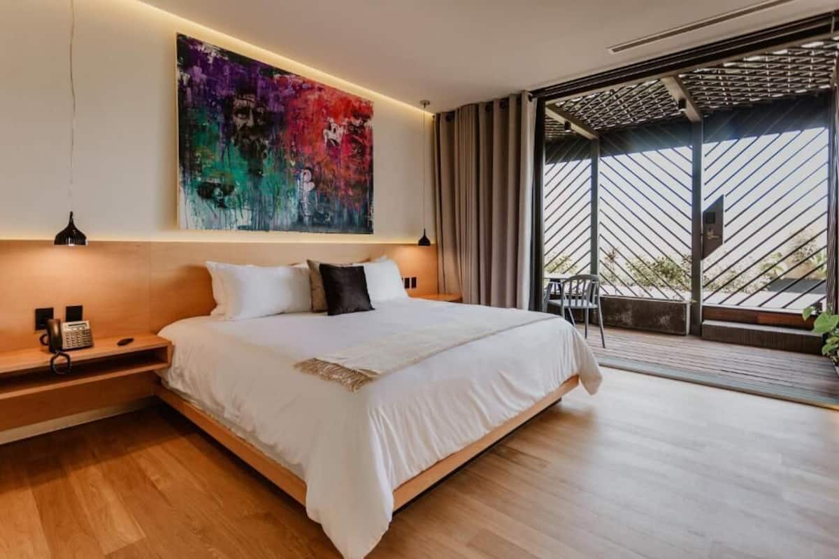 Une chambre moderne avec un grand lit, un téléphone sur la table de chevet, une peinture abstraite aux couleurs vives au-dessus du lit, et une vue sur l’extérieur à travers une grande fenêtre dotée d’une grille métallique.