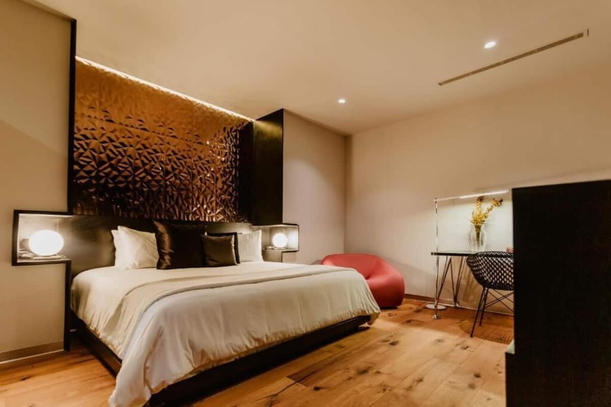 Une chambre à coucher moderne avec un lit, des lampes de chevet, un fauteuil rouge et un bureau agrémenté d’un vase de fleurs.
