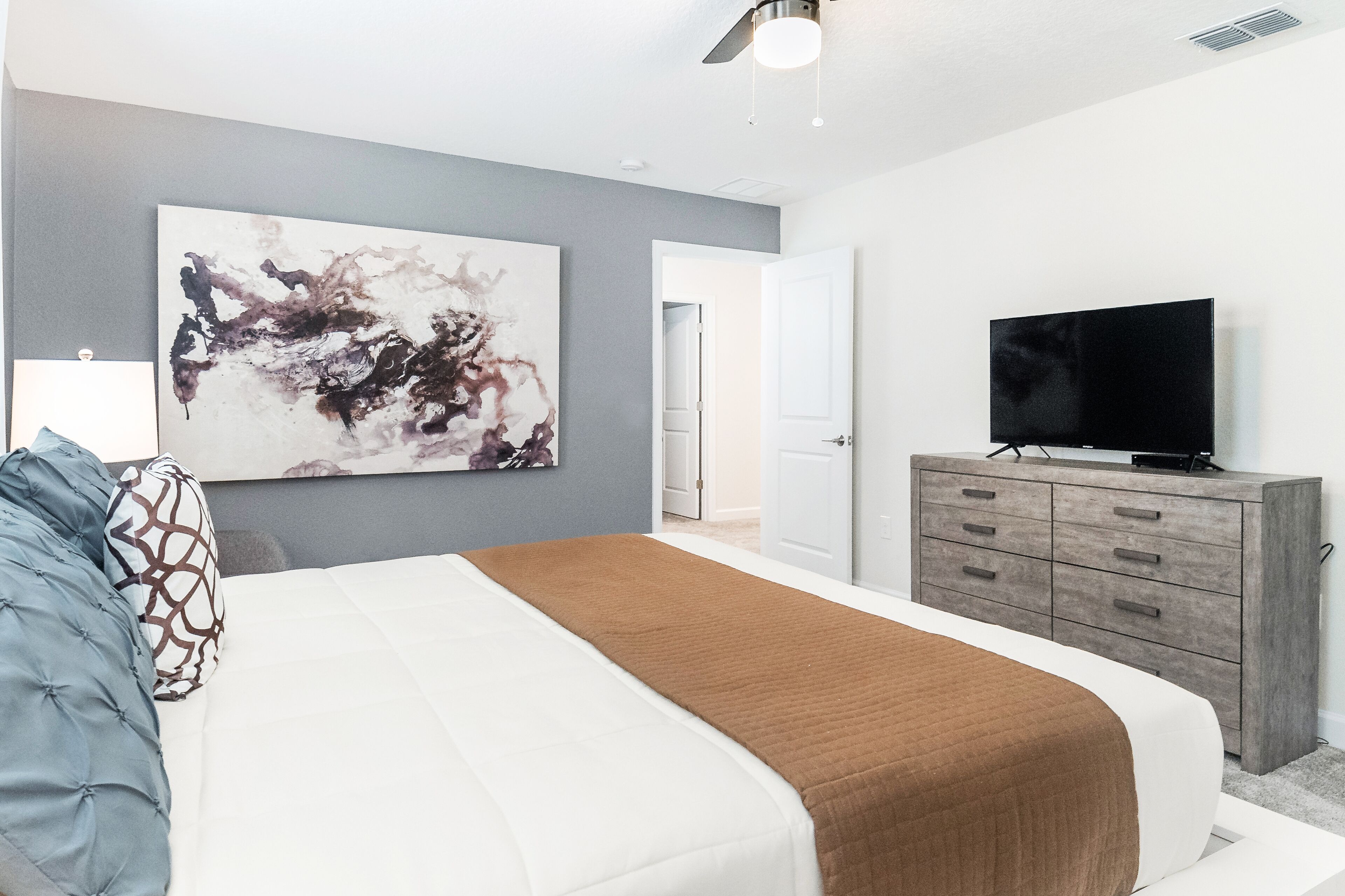 A modern bedroom with a large bed, a flat-screen TV, a gray abstract painting, and a wooden dresser.