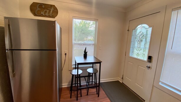 A compact kitchen with a stainless steel refrigerator, a small table with two chairs, a potted plant, and a decorative sign above the refrigerator.