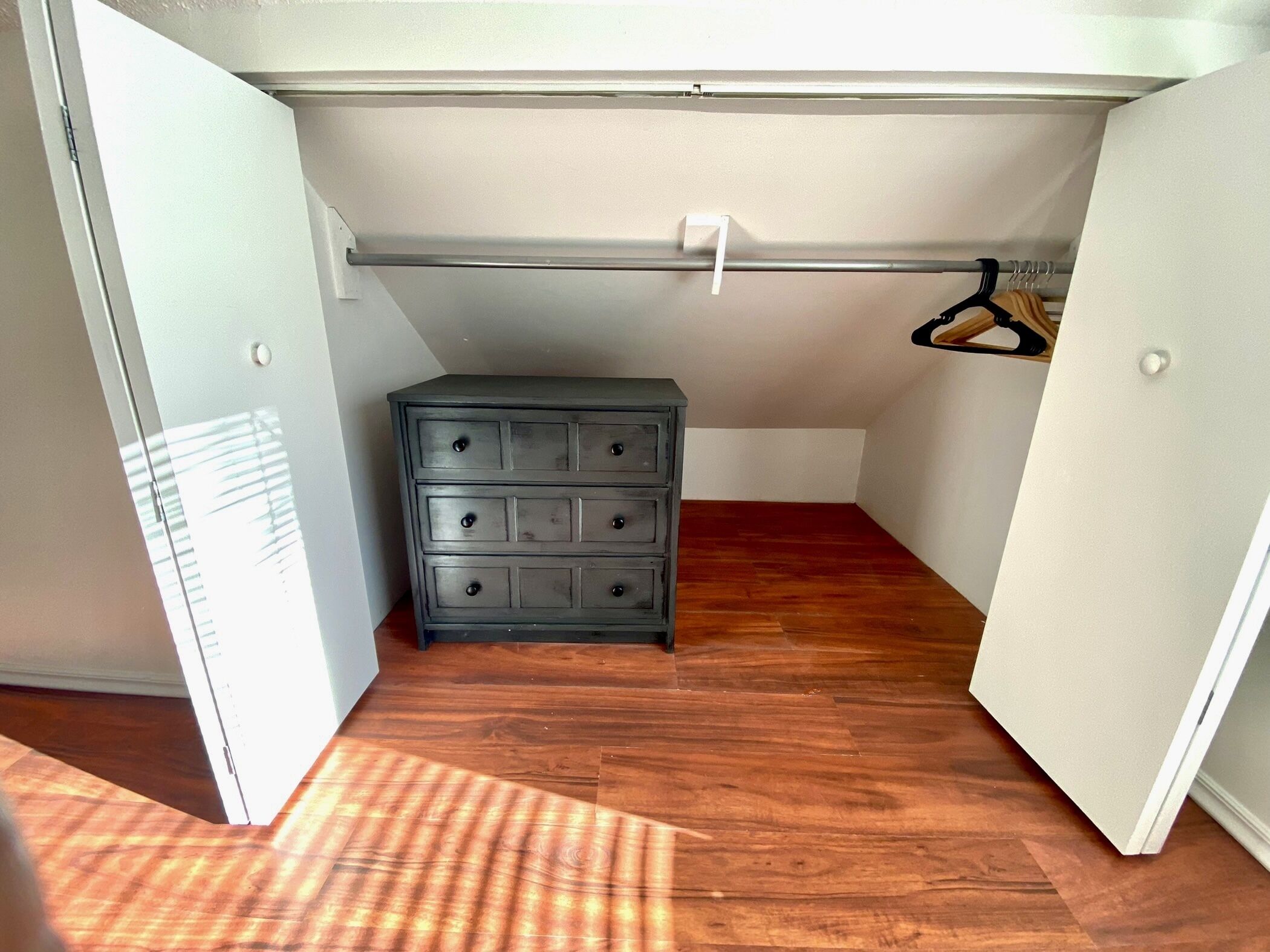 A small, enclosed room with a wooden floor, a gray dresser with multiple drawers, a white ceiling, and a white door.