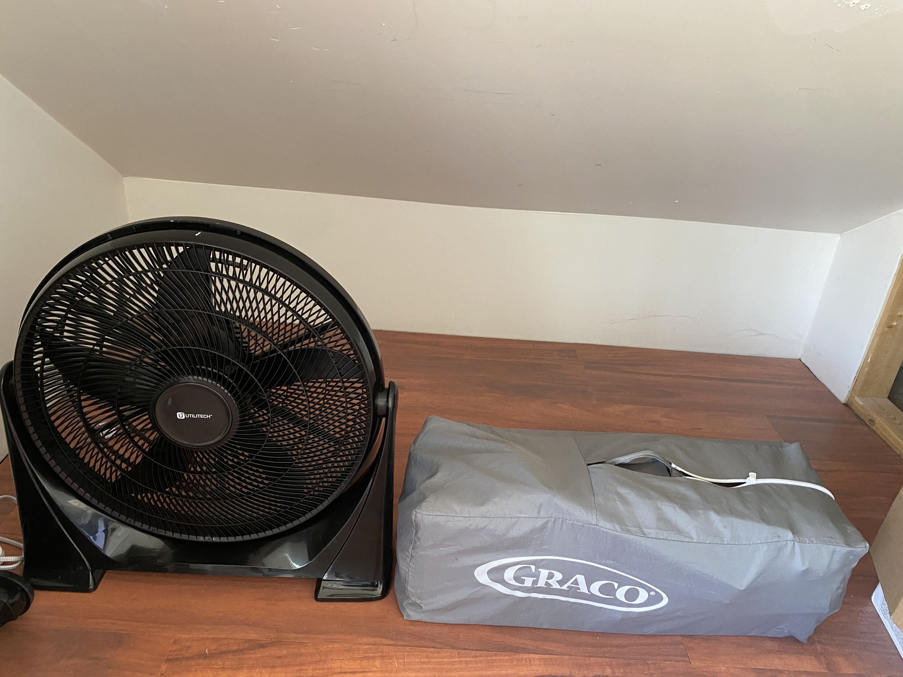 Een zwarte ventilator en een grijze Graco tas op een houten vloer.