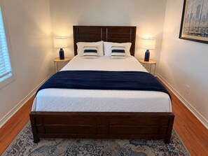 Una cama bien hecha con un cabecero oscuro, dos lámparas de noche y una alfombra estampada sobre un suelo de madera.
