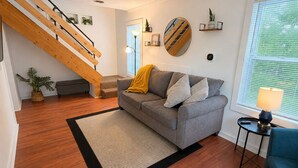A modern living room with a wooden staircase, a grey sofa, a small table with a lamp, and a large window with blinds. - Cozy Lansing Loft - Pet Friendly - Near MSU (Lansing)