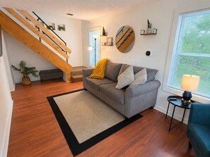 Una sala de estar moderna con una escalera de madera, un sofá gris, una mesita con una lámpara y un ventanal con persianas.