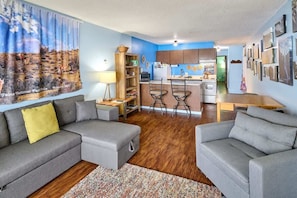 A modern living room with a sofa, a coffee table, and a rug. There is a kitchen area with a refrigerator, a sink, and a dining table.