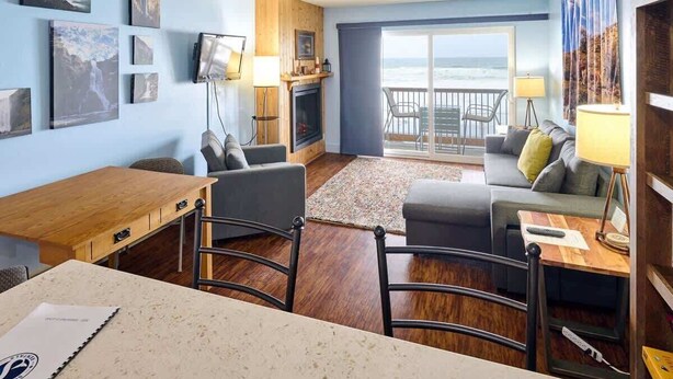 Ein Zimmer mit Holztisch, Stühlen, einer Couch, einem Fernseher, einem Kamin und einem Balkon mit Meerblick.