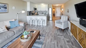 A modern living room with a sofa, coffee table, and TV. A kitchen with a bar and seating area is visible in the background. - Ocean View Condo - Second Floor - Pet Friendly! (Lincoln City)