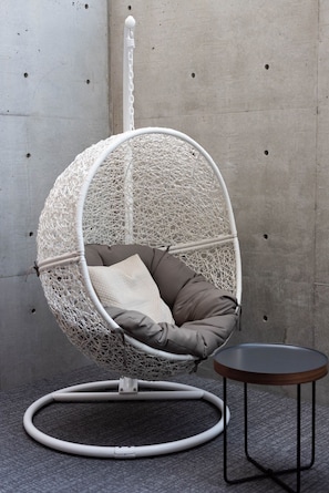 A white wicker hanging chair with a cushion, a small round table, and a cable management system on a gray carpet.