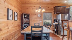 Ein Holzblockhaus-Interieur mit Esstisch, Stühlen, einer Deckenleuchte, einer Uhr und einem Kühlschrank.