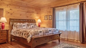Ein Schlafzimmer einer Holzhütte mit einem Bett, zwei Nachttischlampen, einem Nachttisch und einem Fenster mit Vorhängen.