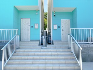 Un escalier menant à un bâtiment doté de deux portes, chacune étant marquée d’une lettre, et des bouteilles de gaz à l’extérieur.