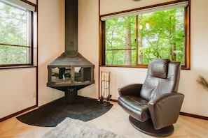 A cozy room with a fireplace, a leather chair, and large windows offering a view of trees.
