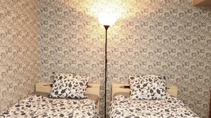 Une chambre avec deux lits, chacun recouvert d’une couvre-lit à motifs floraux et agrémenté d’oreillers assortis. Une lampe de sol à pied noir et abat-jour blanc est placée entre les lits. Les murs sont recouverts d’un papier peint à motifs floraux en relief.