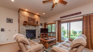 Una sala de estar acogedora con chimenea de piedra, sofás color beige cómodos, un televisor de pantalla plana y un amplio ventanal con vistas al exterior.
