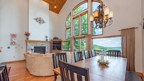 Un comedor amplio con una gran mesa de madera y sillas, chimenea y ventanas grandes que ofrecen vistas al exterior.