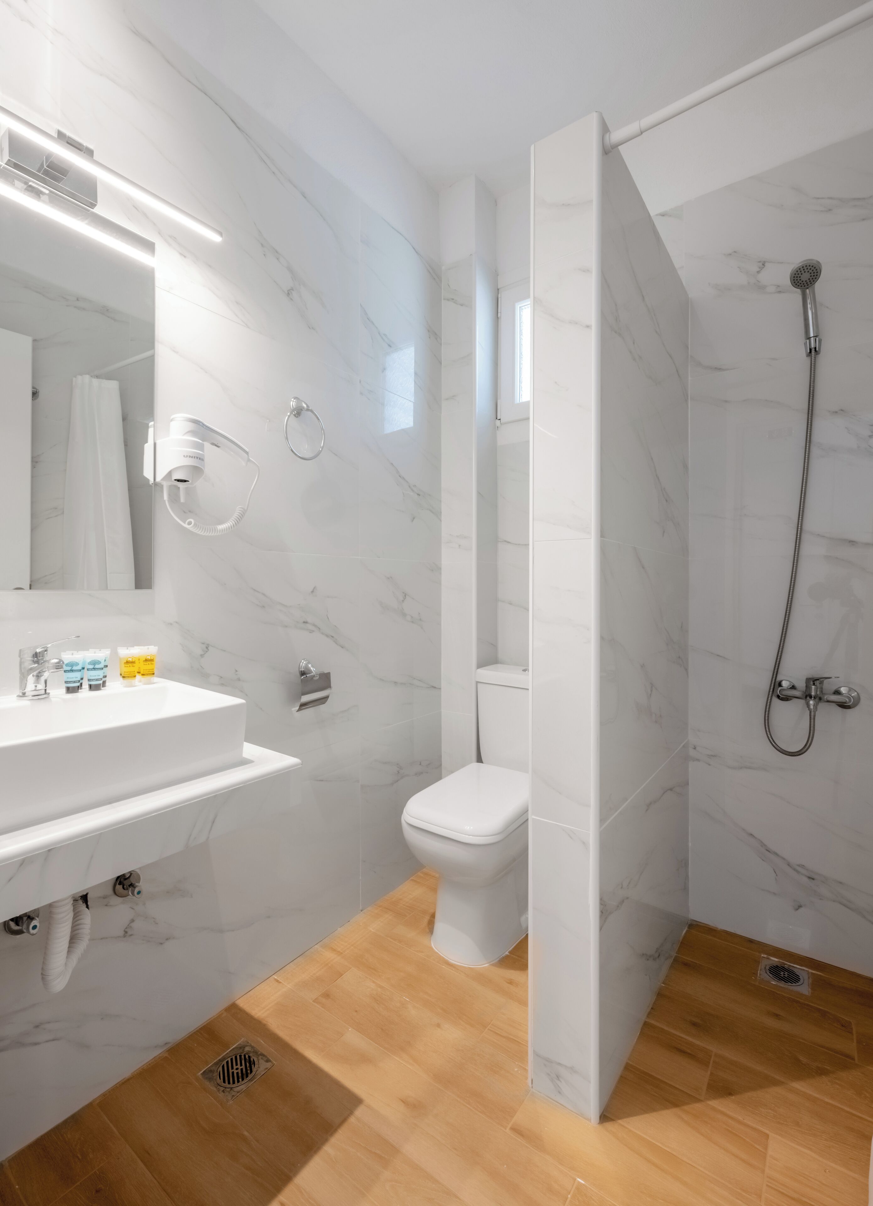 Een moderne badkamer met een marmeren vloer, een witte wastafel met toiletartikelen, een toilet en een douchehoek.