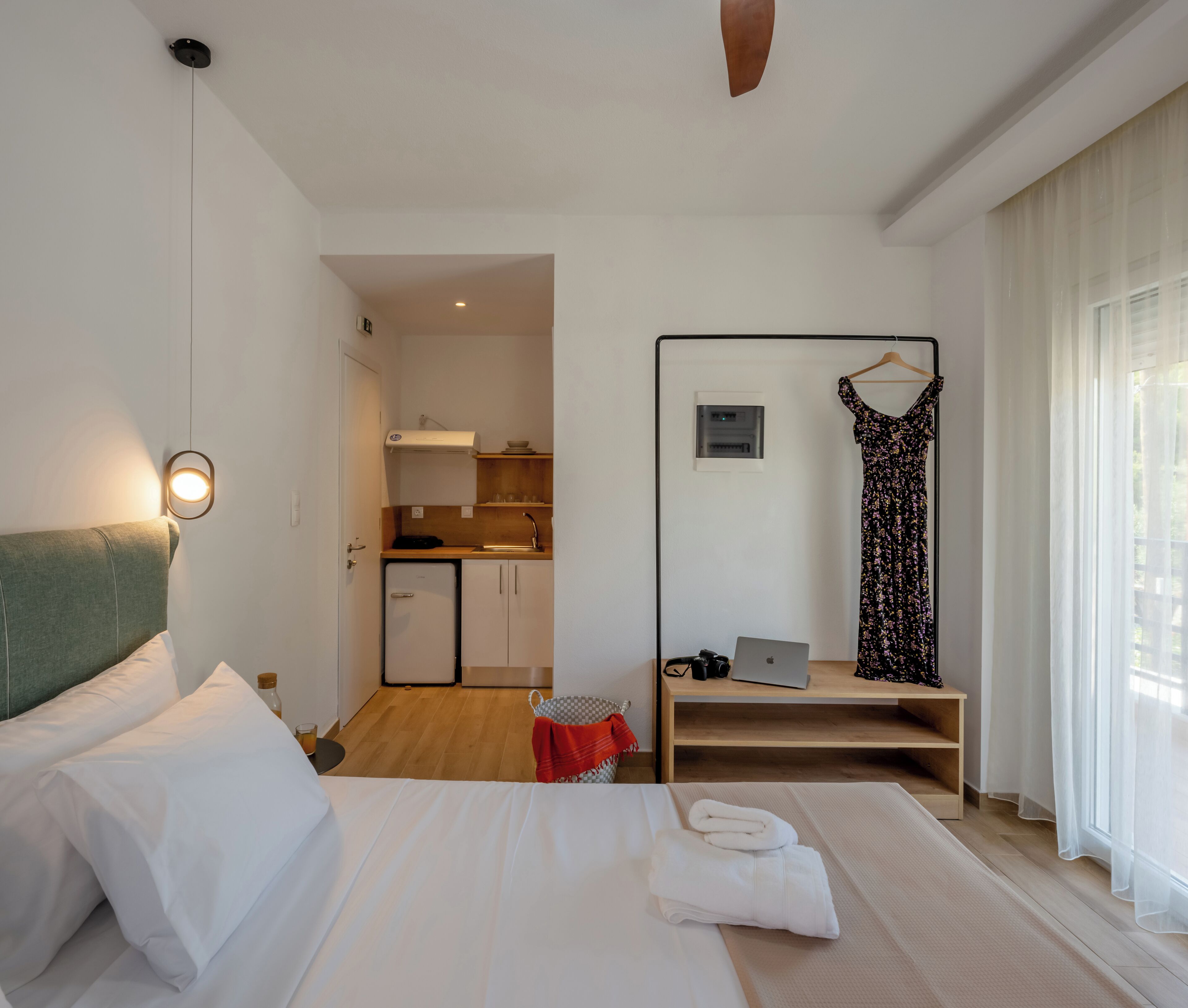 Habitación de hotel con cama, un escritorio con una computadora portátil, un vestido colgado y una kitchenette al fondo.