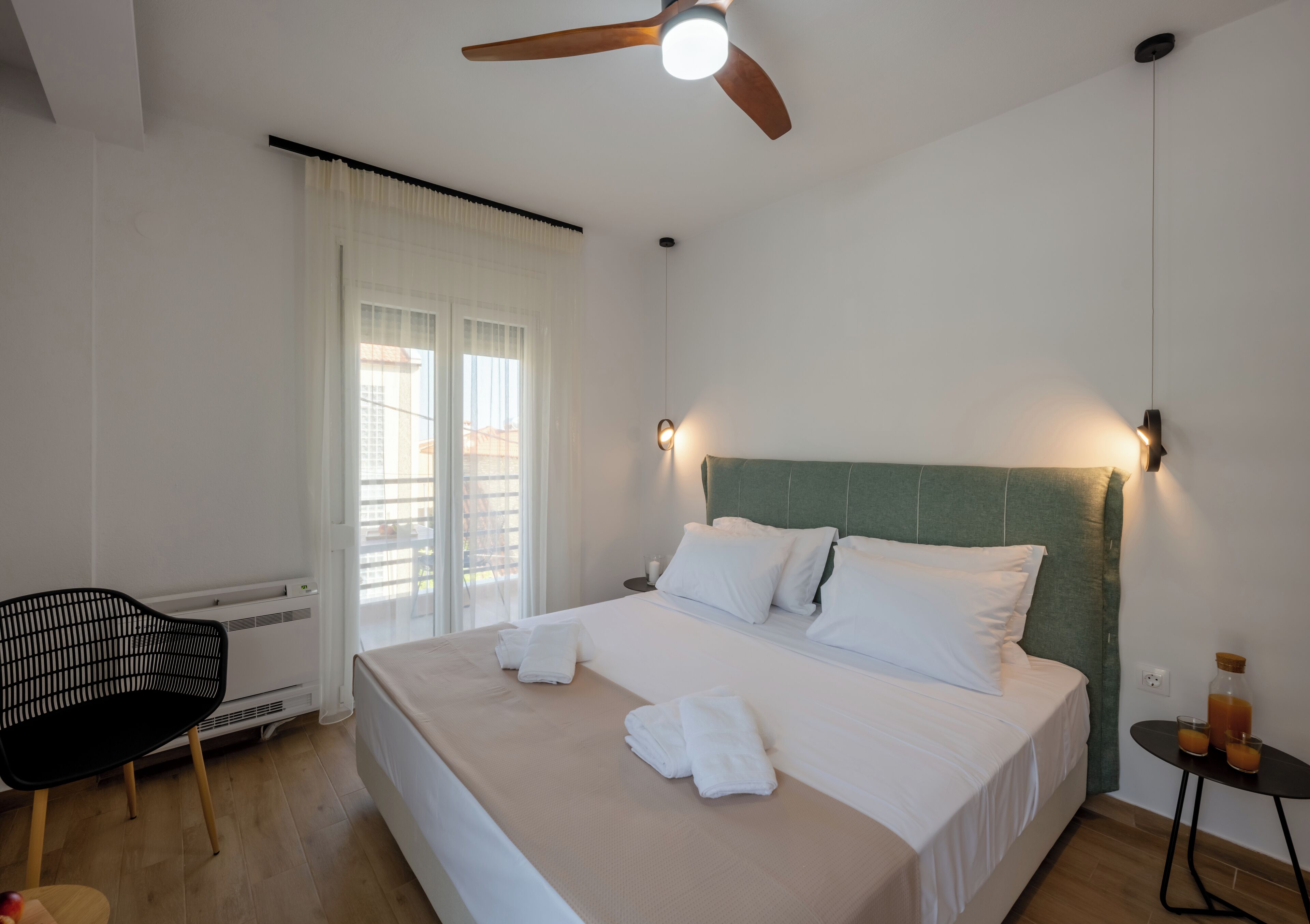 Quarto de hotel com uma cama, uma cadeira, uma mesa com garrafa e copos, e um ventilador de teto.