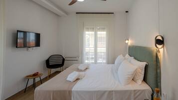 Una camera d'albergo con un letto, una ventola a soffitto, una TV, una sedia, un tavolino e una finestra con tende.