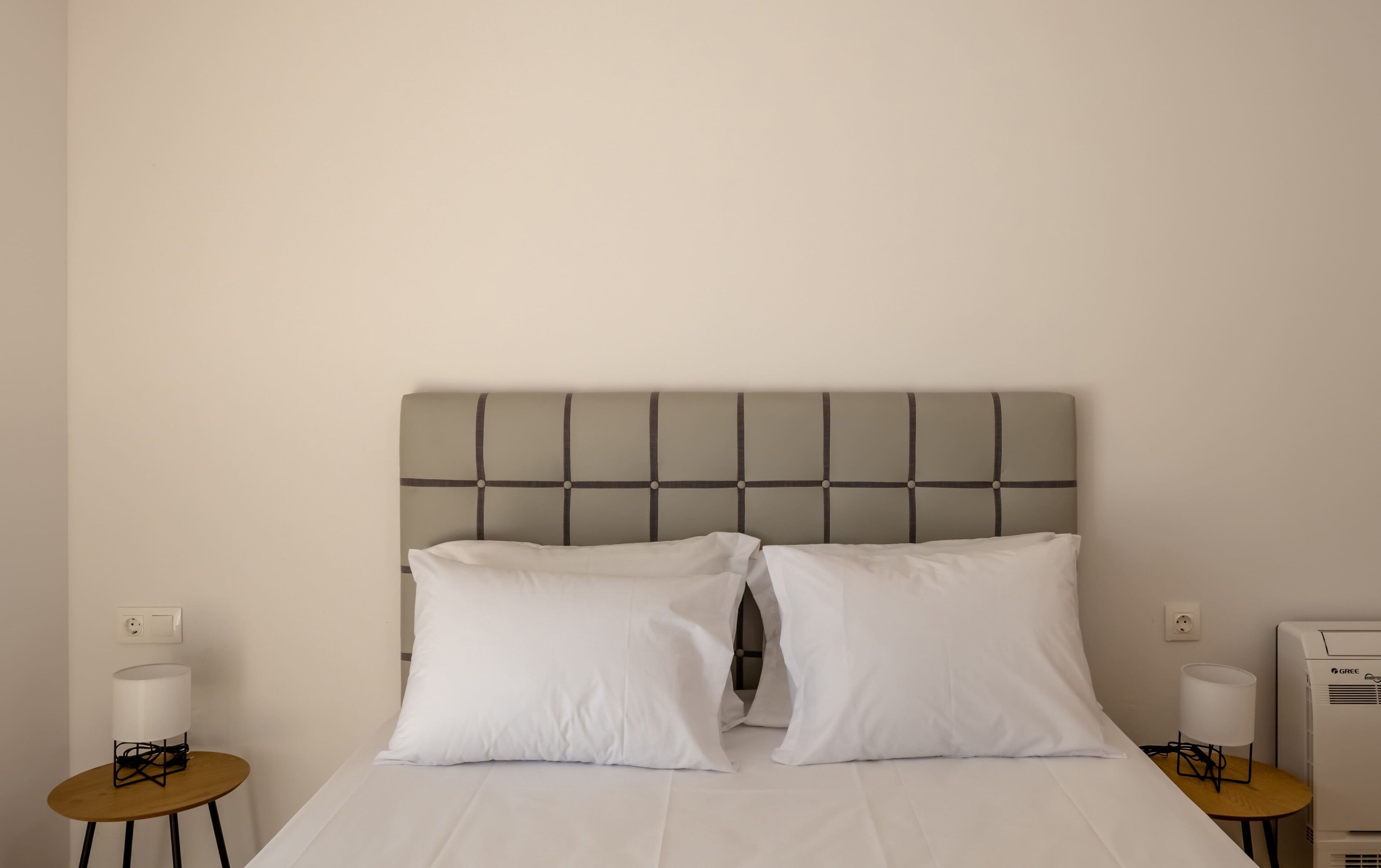 Una cama bien hecha con un cabecero gris, dos almohadas blancas y una mesita de noche con una lámpara.