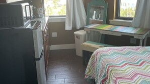 Cocina compacta con refrigerador, microondas y una pequeña mesa de comedor. Hay una ventana con cortinas, una cama con un edredón colorido y un espejo en la pared.